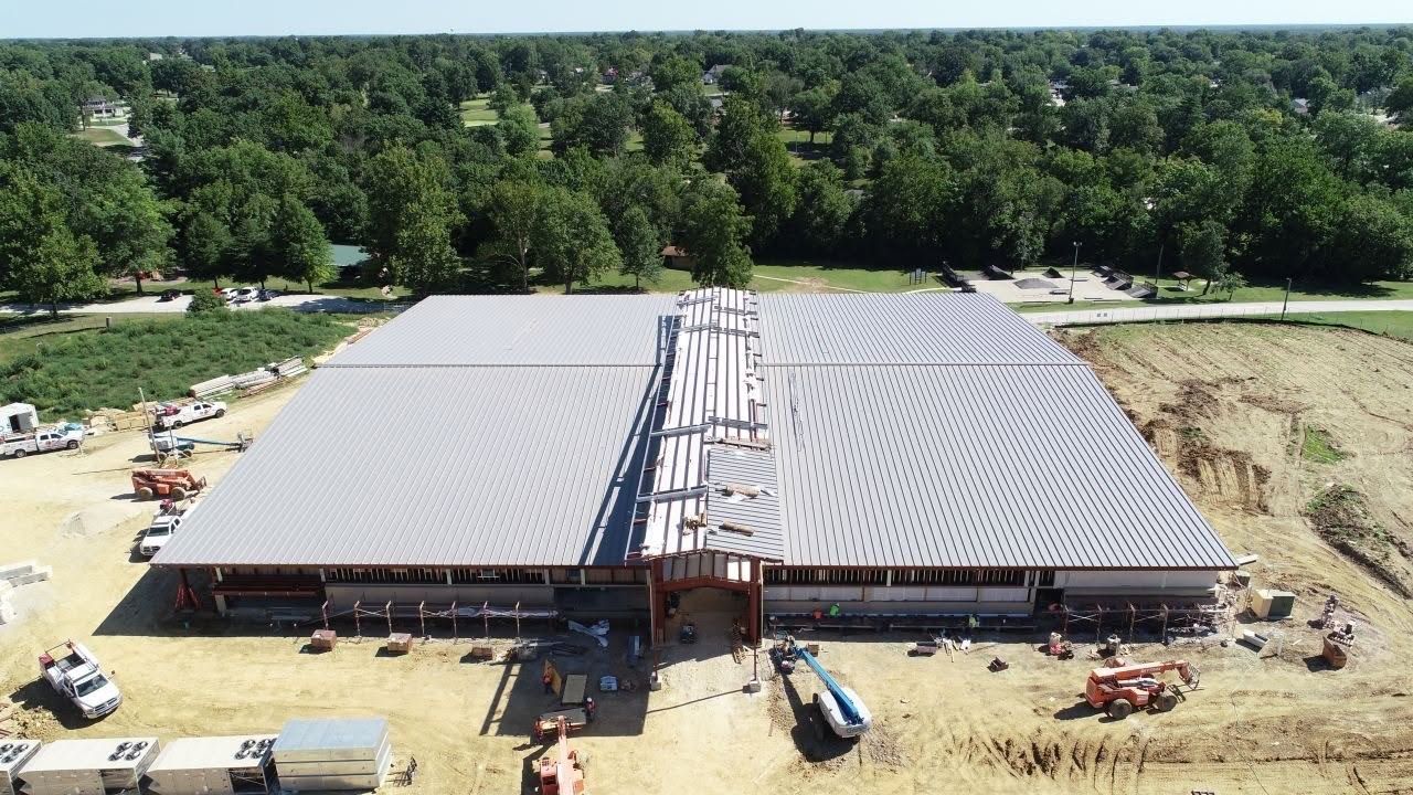 Aerial view of a large building with its metal roof panels installed and crews working along the wall line